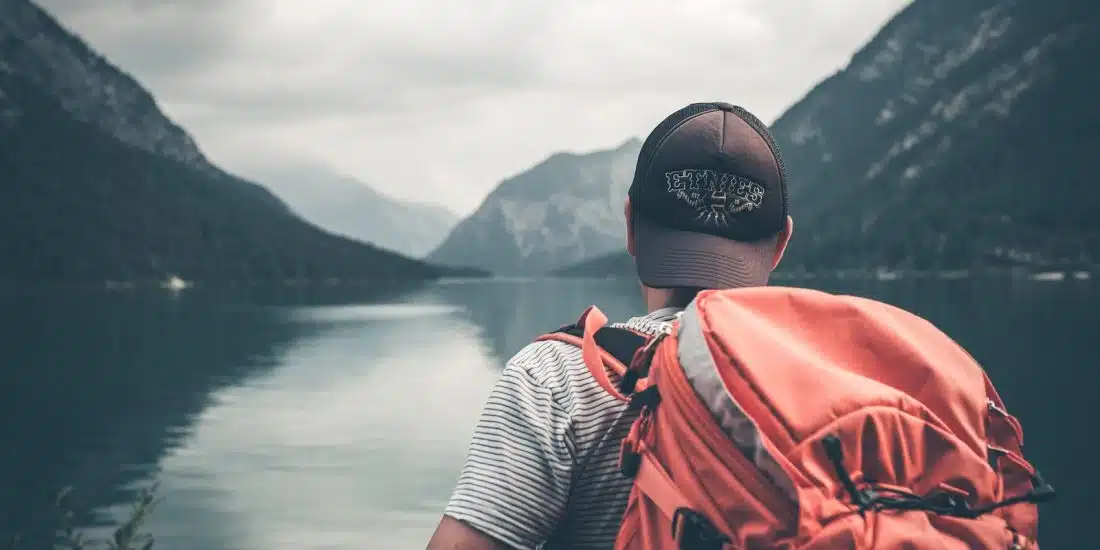 man with red hiking backpack facing body of water and mountains at daytime
